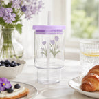 Floraler Glasbecher mit lila Deckel und Strohhalm in heller Tischszene